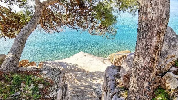 Un escalier en pierre mène à une plage bordée d’une eau d’un bleu cristallin.