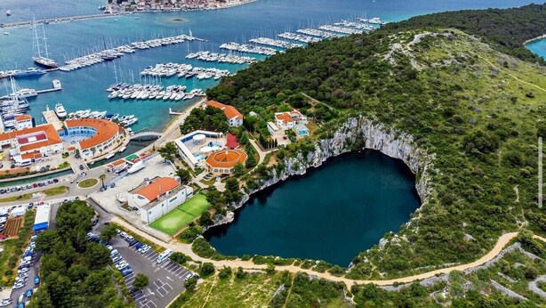 Vista aérea de una zona costera con un puerto deportivo, un acantilado con un lago y un conjunto de edificios con techo rojo.