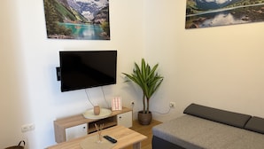 Una sala de estar con un televisor de pantalla plana, una mesa de centro de madera, un sofá gris, una planta en maceta y dos cuadros de paisajes en la pared.