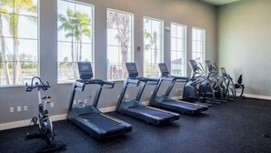 Et fitnessrum med løbebånd og crosstrainer, store vinduer med udsigt til palmer.
