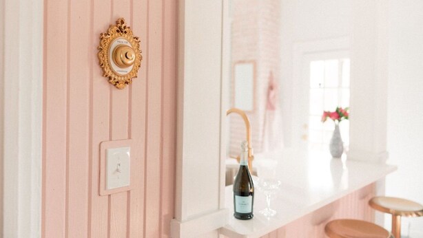 A bathroom with a pink wall, a gold-framed mirror, a white light switch, a bottle of wine, and a glass on a white countertop.
