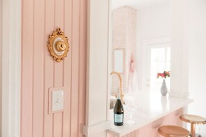 Un baño con una pared rosa, un espejo con marco dorado, un interruptor de luz blanco, una botella de vino y una copa sobre una encimera blanca.