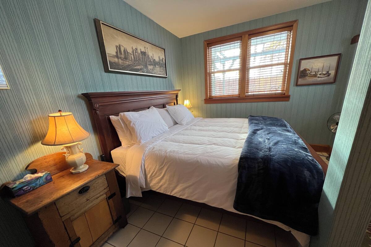 Una cama bien tendida con sábanas blancas, una mesita de noche de madera con una lámpara, y una ventana con persianas.