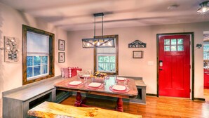 Comedor con mesa de madera dispuesta para cuatro, una puerta roja y una ventana con persianas.