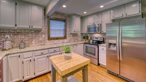 Una cocina con armarios blancos, pared de ladrillo, mesa de madera con una planta, electrodomésticos de acero inoxidable y una ventana con persianas.