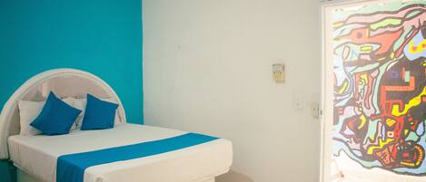 A bed with a white headboard and blue pillows, a blue accent on the bedspread, and a colorful abstract painting on the wall.
