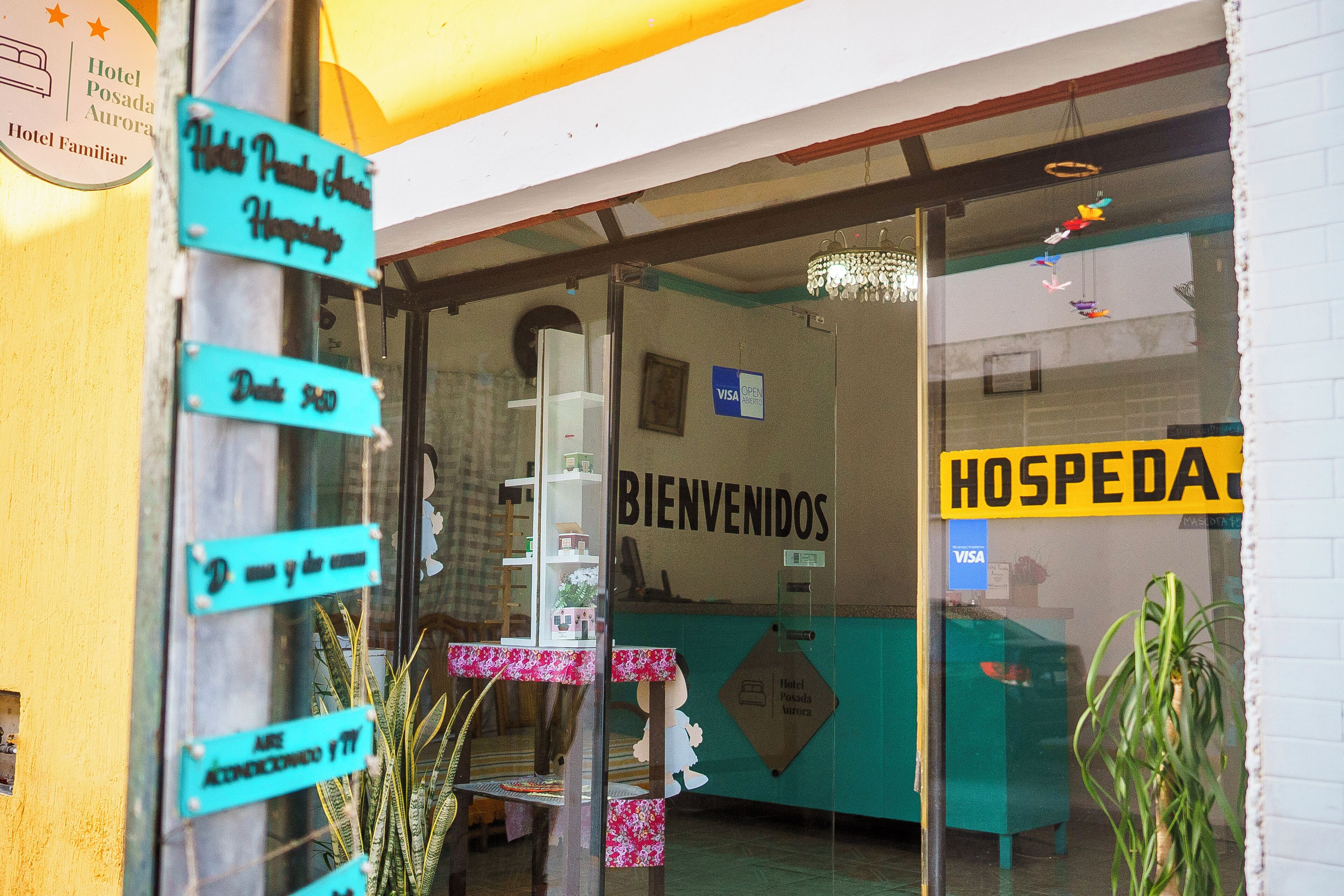 Zona de recepción de hotel con un cartel que dice 'BIENVENIDOS', un cartel de 'VISA' y un cartel de 'Hotel Posada Aurora'.