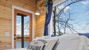 Een houten slaapkamer met een bed, een kussen met tekst, een raam en uitzicht op bomen.