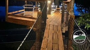Een houten terras met een nethek, omringd door bomen en lichtjes, met uitzicht op een wateroppervlak.