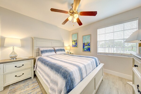 A bedroom with a bed, nightstands, a ceiling fan, artwork, and a window with blinds.