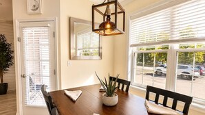 Un comedor con una mesa de madera, sillas, lámpara colgante, planta en maceta y ventana con persianas.