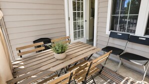 Un patio exterior con una mesa y sillas de madera, una planta en maceta y vista del interior de la casa a través de una puerta.