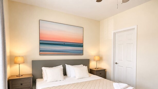 A bedroom with a bed, bedside tables, a wooden table, a door, and a painting on the wall. - Surf Shack Condo! Centrally located to beaches, shopping, and entertainment (Rehoboth Beach)
