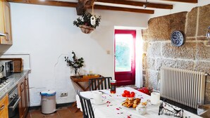 A cozy kitchen with a dining table set for four, featuring a white tablecloth with a cherry pattern. There is a stone wall, a window with a red frame, and a hanging plant.