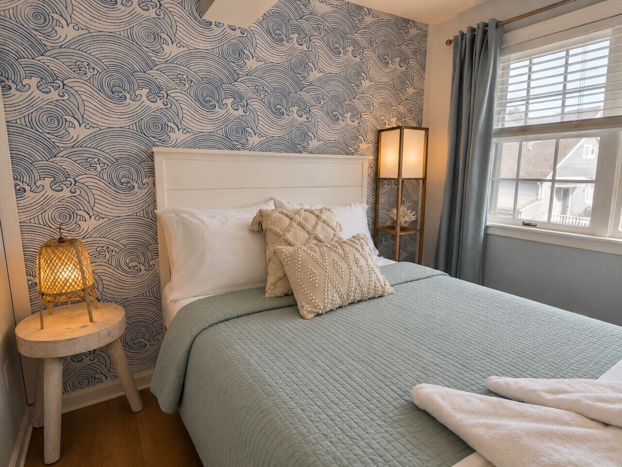 A bedroom with a patterned wallpaper, a bed with a quilted bedspread, two bedside lamps, and a window with curtains.