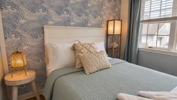 A bedroom with a patterned wallpaper, a bed with a quilted bedspread, two bedside lamps, and a window with curtains.