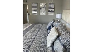 A bedroom with a patterned bedspread, three framed artworks on the wall, and a bedside lamp.