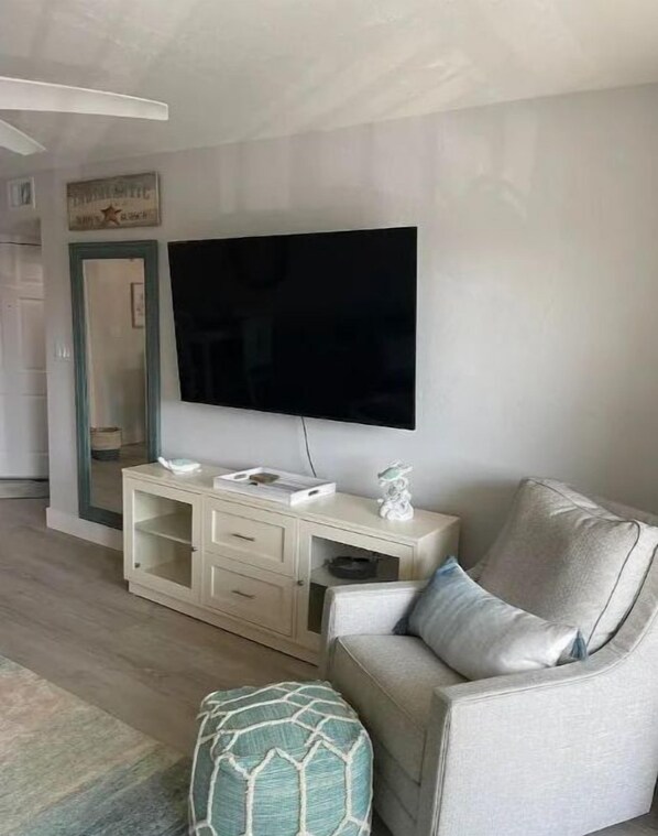 Un salon doté d’un téléviseur à écran plat fixé au mur, d’un canapé de couleur claire, d’un pouf assorti et d’une console blanche agrémentée d’objets décoratifs.