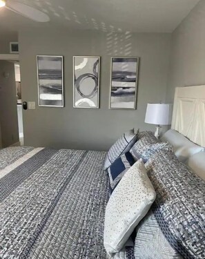 Une chambre avec une couvre-lit à motifs, trois œuvres d’art encadrées accrochées au mur et une lampe de chevet.