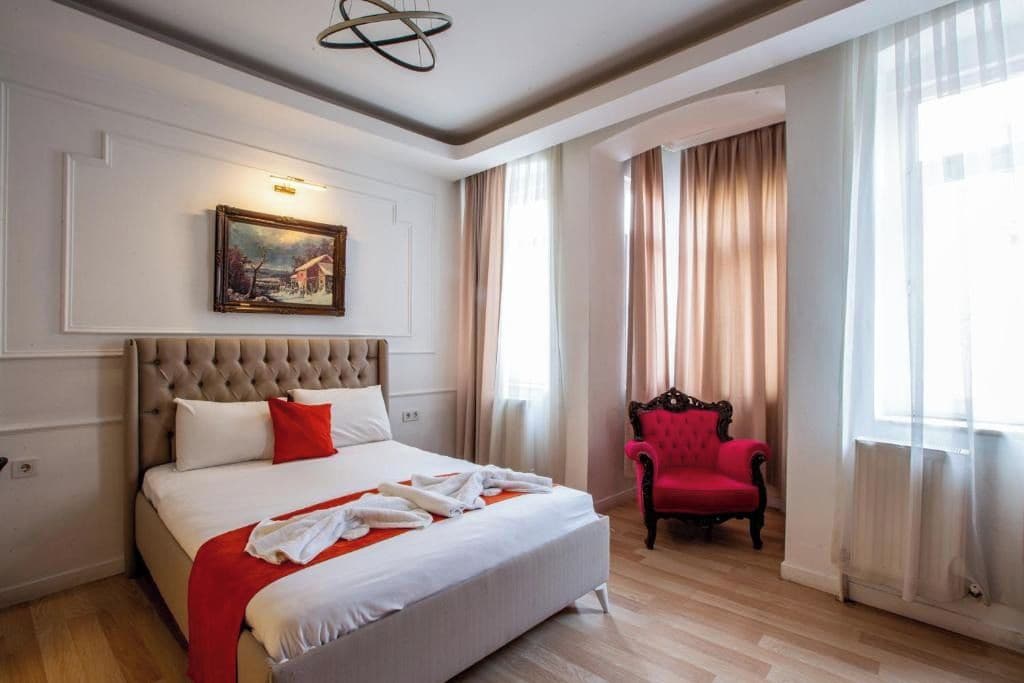 A hotel room with a large bed, a red armchair, a painting on the wall, and a window with curtains.