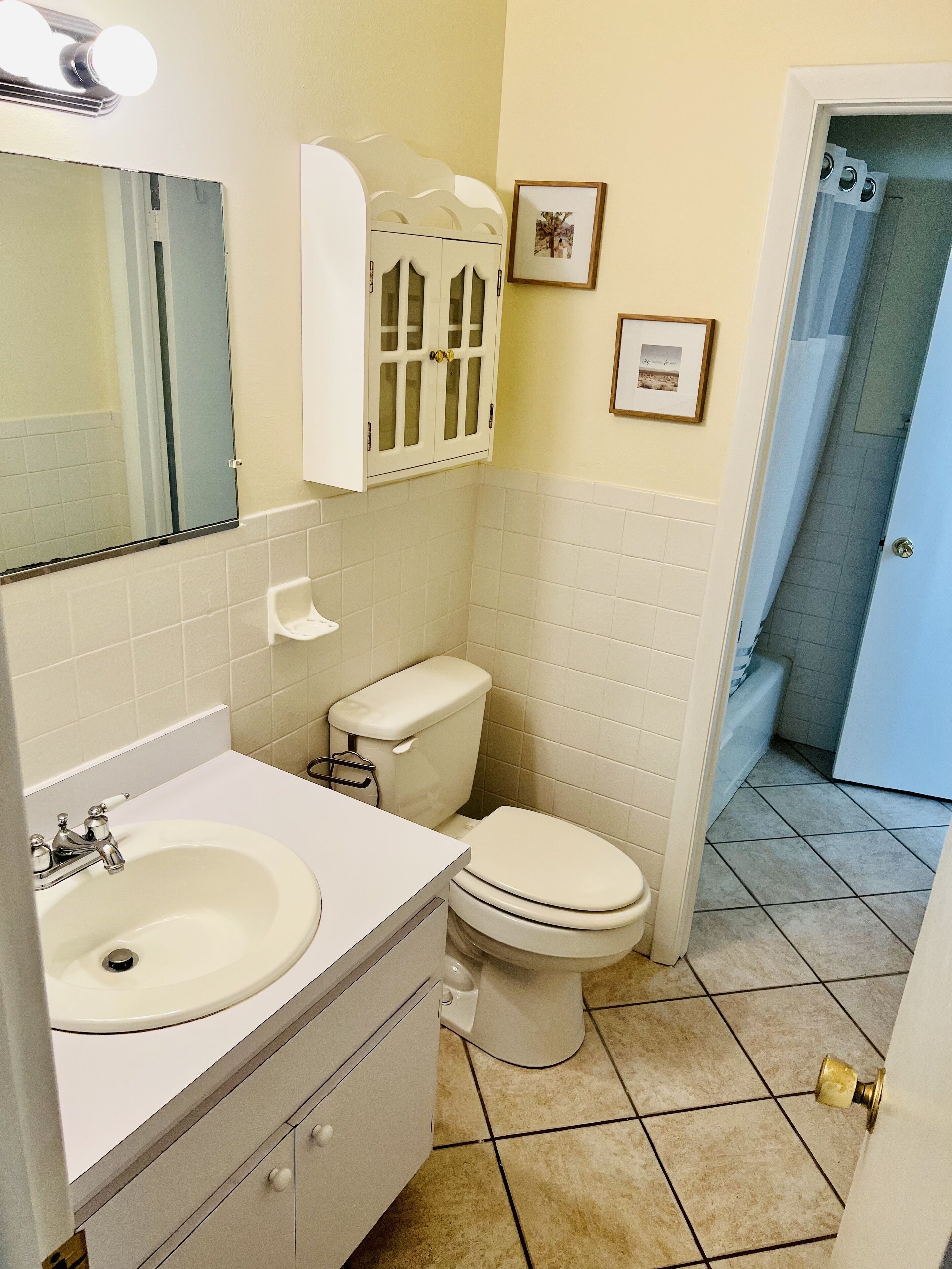 Een badkamer met een witte wastafel, toilet en een badkamermeubel. Twee ingekaderde foto's hangen aan de muur en er is een douchegordijn zichtbaar op de achtergrond.