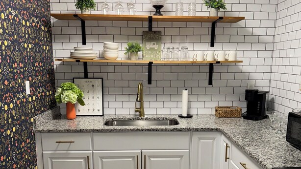 A modern kitchen with white cabinets, a granite countertop, and a black stove. There are two shelves above the sink holding glassware and plants. - Sweet Magnolias Home: Walk to Square (Covington)