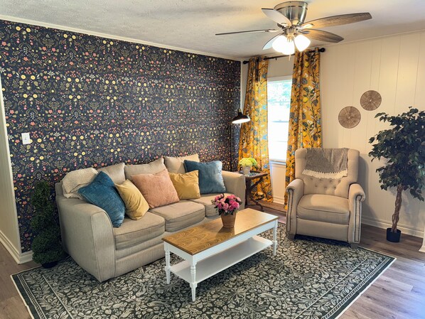 Ein Wohnzimmer mit blumengemusterten Wand, einem Deckenventilator, einer Couch mit Kissen, einem Couchtisch und einem Sessel.