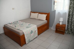 Un lit simple avec une couvre-lit à motifs floraux, une table de chevet en bois avec une lampe, et une fenêtre ornée de rideaux légers.