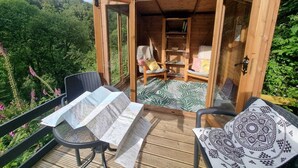 A wooden cabin with a balcony, a chair, a table, and a bookshelf. - Cosy Cottage overlooking the Teifi Gorge (Cilgerran)