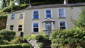 A two-story house with a front porch, a small garden, and a stone staircase leading to the entrance. - Cosy Cottage overlooking the Teifi Gorge (Cilgerran)