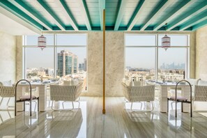 A modern dining area with a city view, featuring a central column, white furniture, and hanging lanterns. - Abesq Doha Hotel and Residences - An IHG Hotel (Doha)