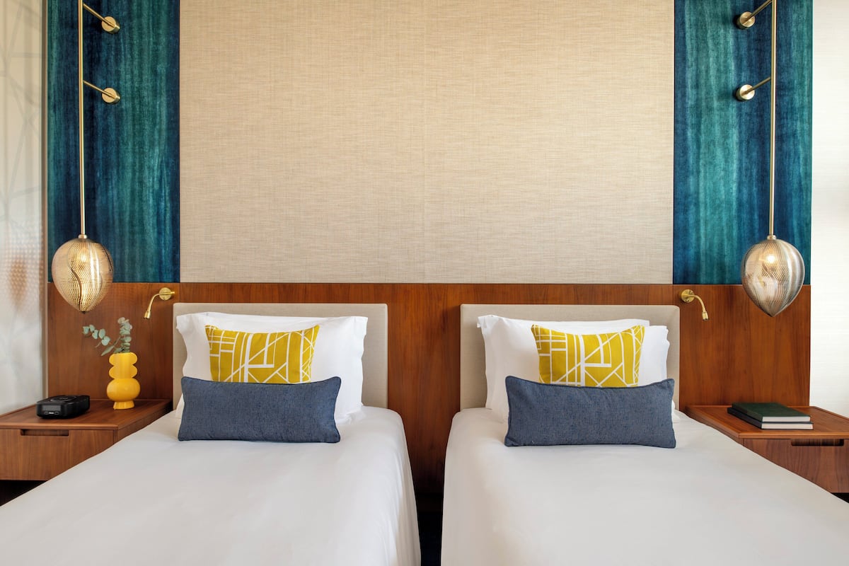 Habitación de hotel con dos camas, cabeceros de madera y una pared en tonos neutros.