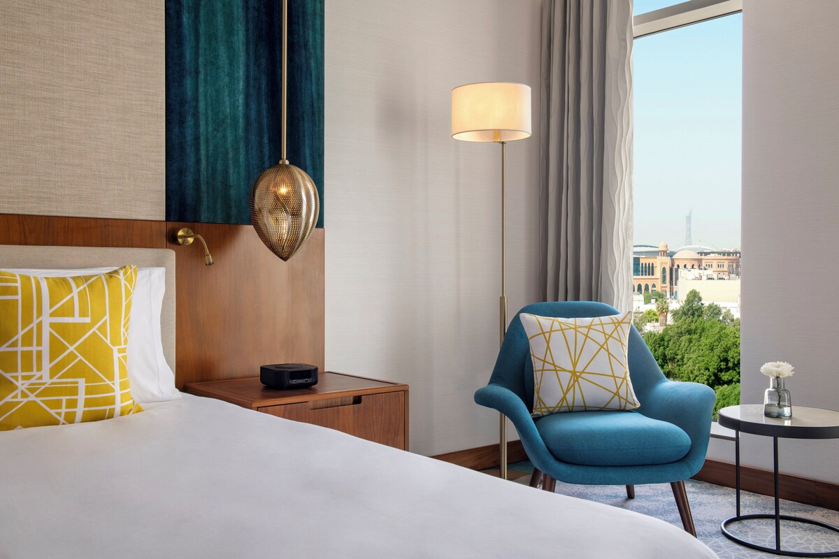 A hotel room with a bed, a blue armchair, a small table, and a view of buildings through the window.