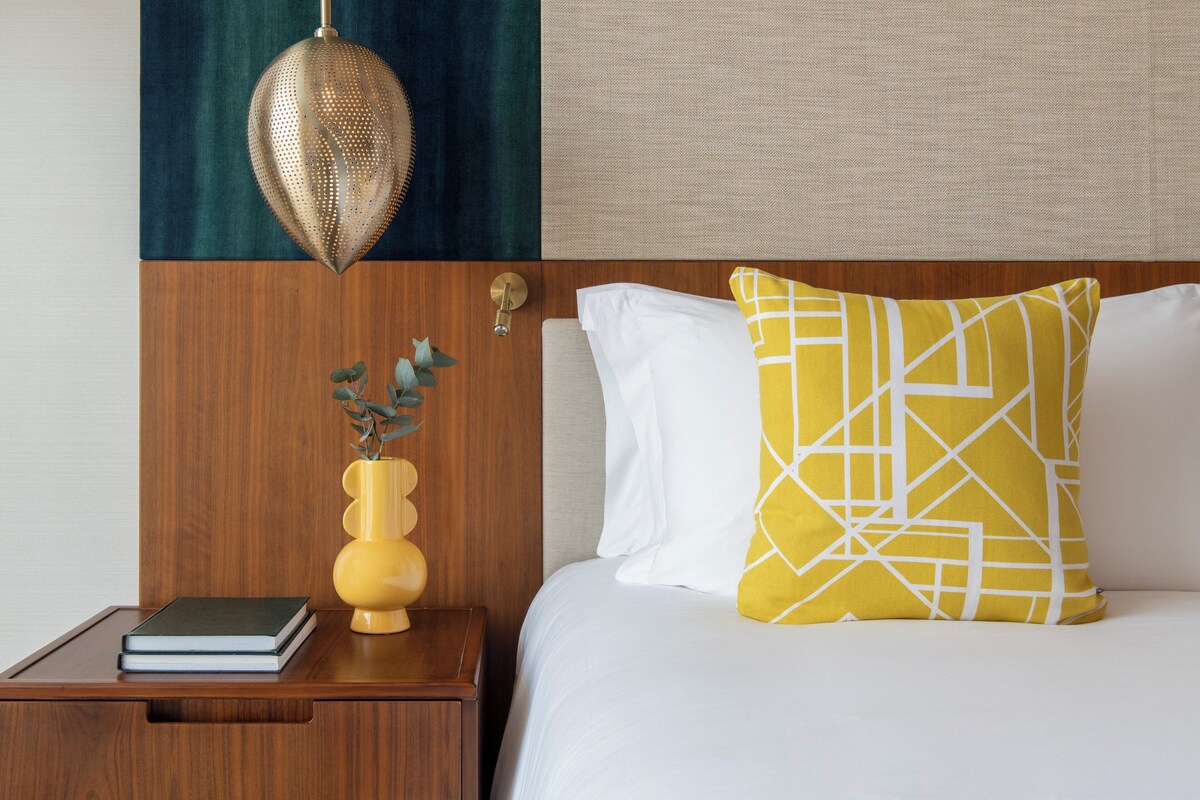 A neatly made bed with white and yellow pillows, a wooden nightstand with a book and a yellow vase with green leaves, and a hanging light fixture.