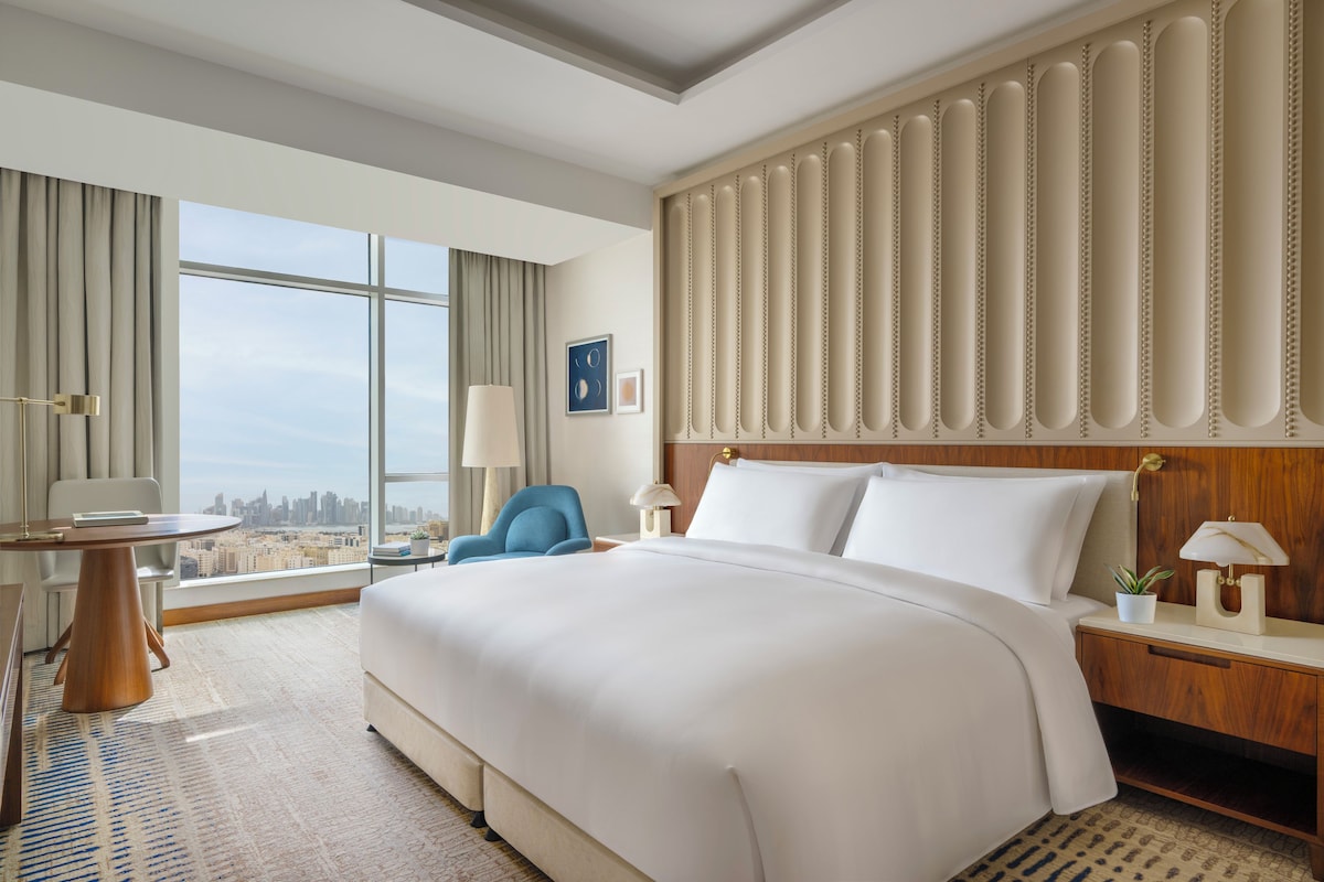 A hotel room with a large bed, a desk, a chair, and a view of the city.
