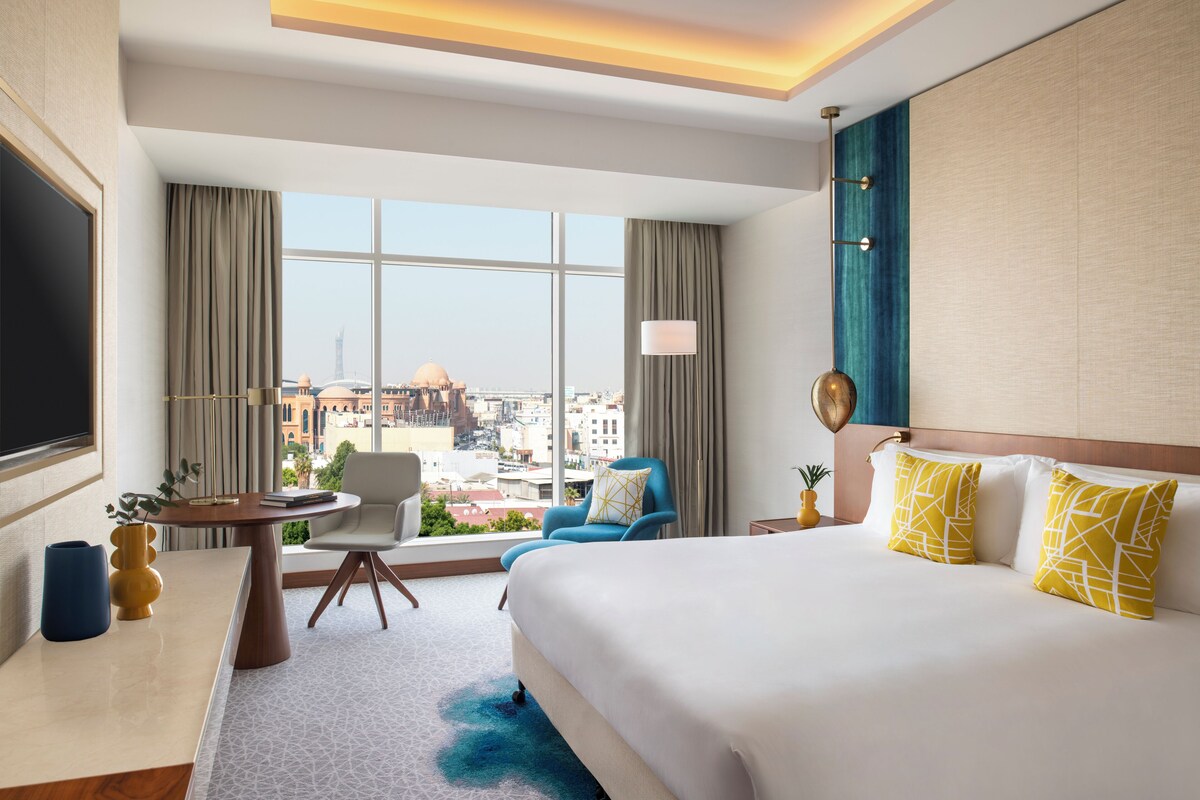 A hotel room with a large bed, a desk, a chair, and a view of the city.