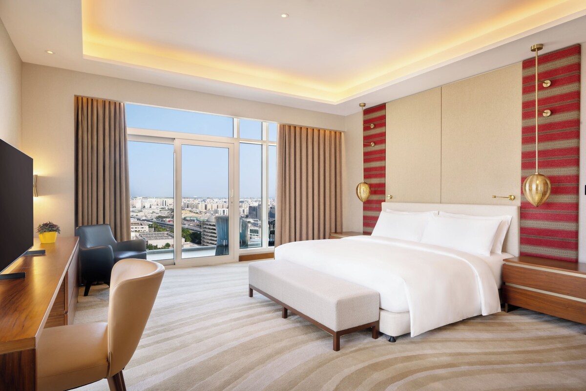 A hotel room with a large bed, a desk, a chair, and a view of the city.