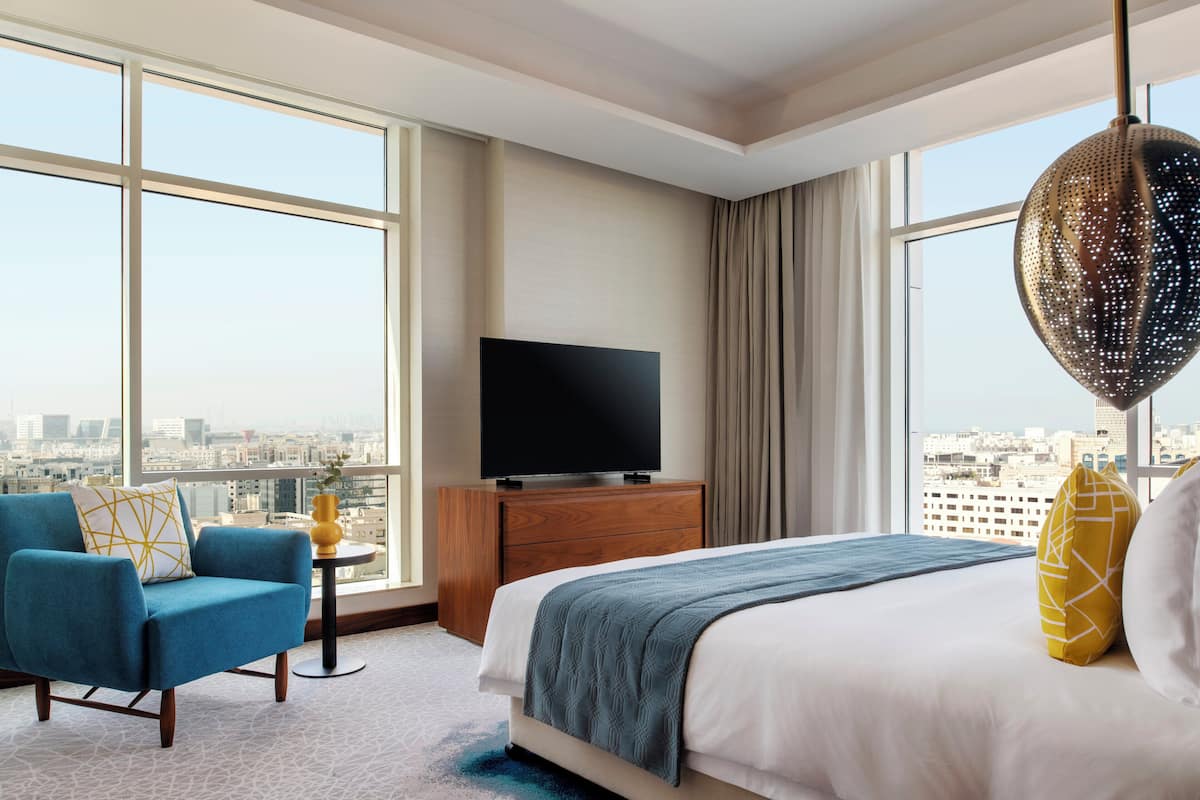 Habitación de hotel con una cama grande, televisión, una silla y vista a la ciudad.