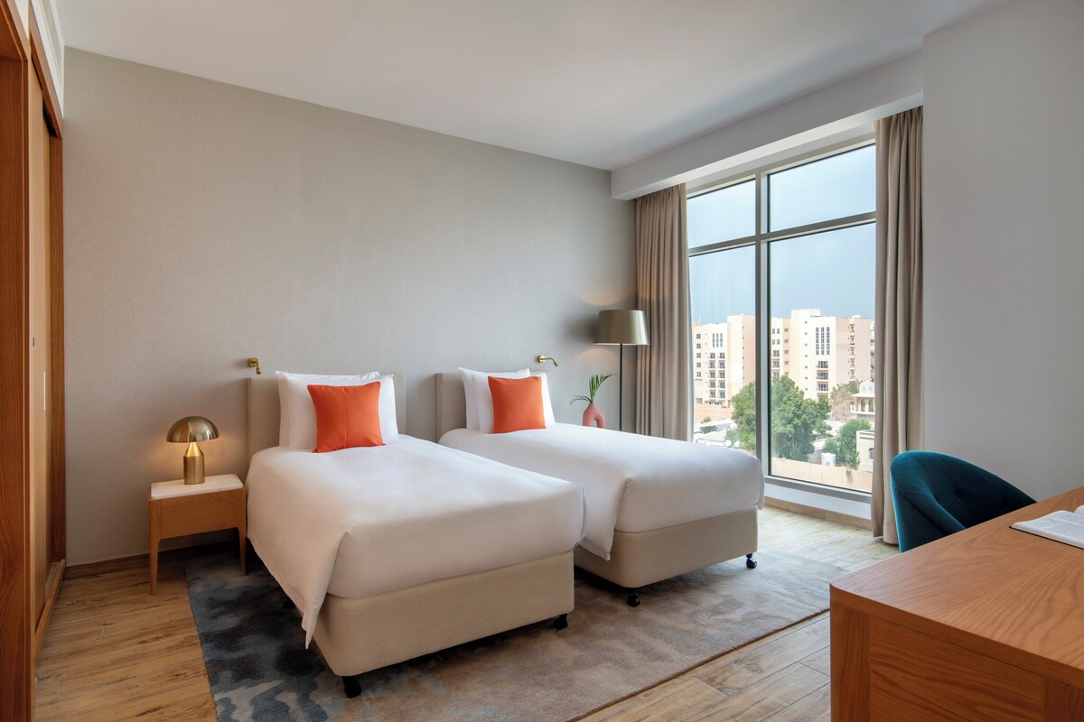 A hotel room with two beds, a desk, a lamp, and a view of buildings through large windows.