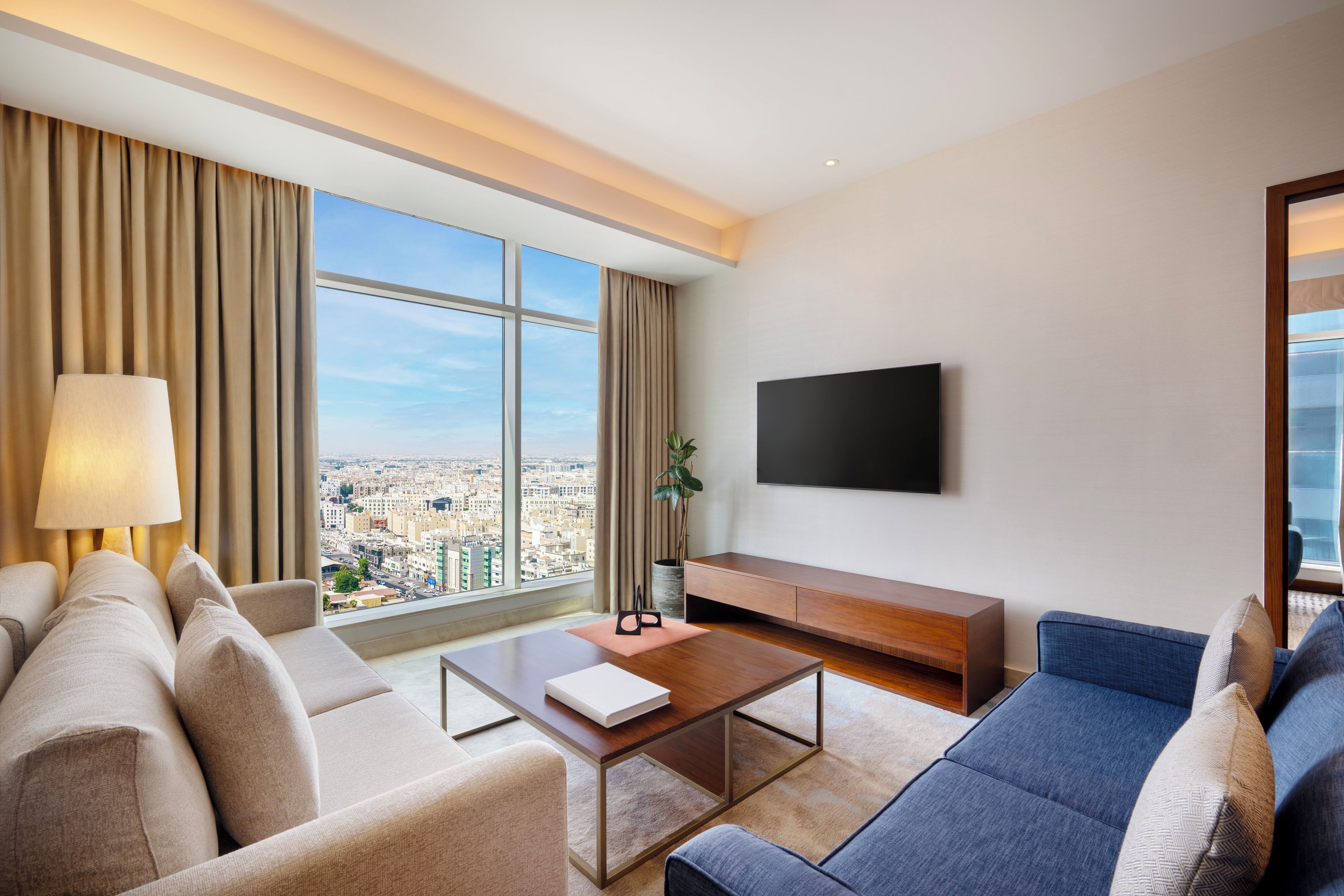 A modern hotel room with a city view, a flat-screen TV, a sofa, a coffee table, and a lamp.