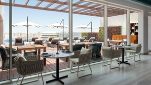 Un área de comedor exterior moderna con amplios ventanales, sombrillas y vistas a la ciudad.