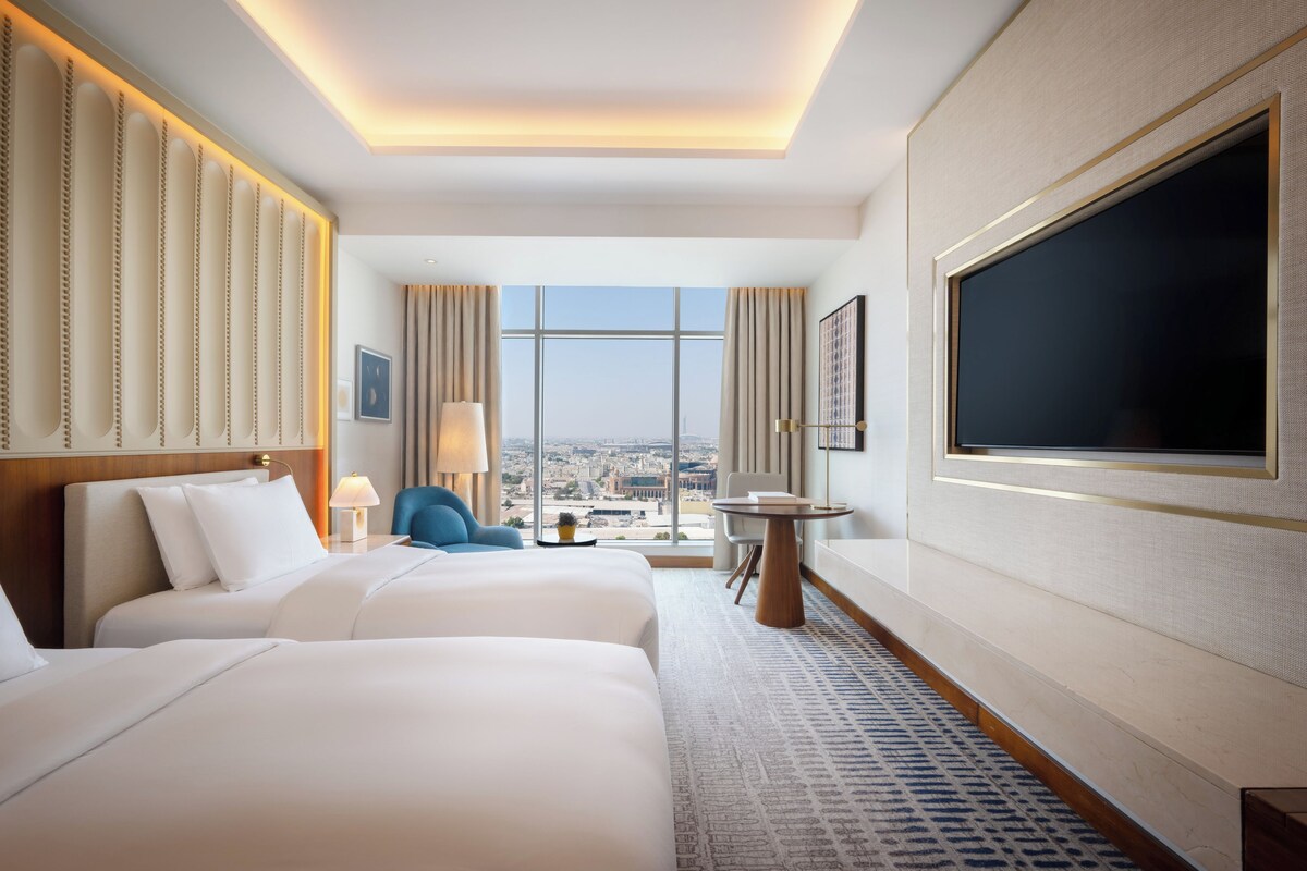 A hotel room with two beds, a large TV, a city view, and a modern design.