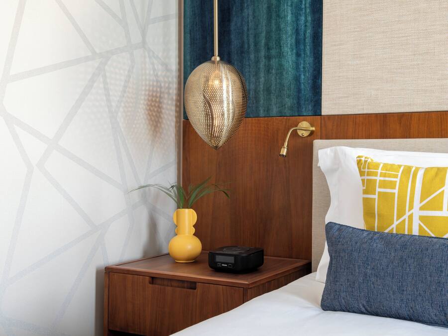 A neatly made bed with a wooden headboard, a bedside table with a small plant and a black device, and a hanging light fixture.