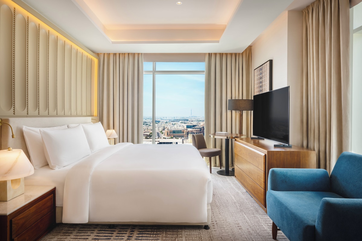 A hotel room with a large bed, a television, a desk, and a view of the city.
