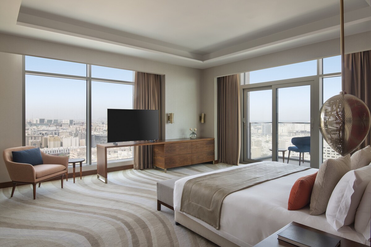 A modern hotel room with a large bed, a desk with a TV, a sofa, and a city view through large windows.