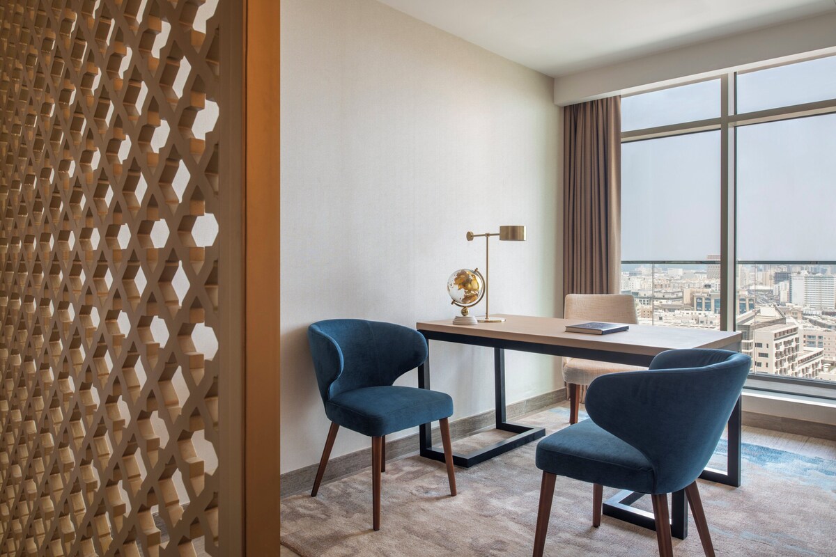 A modern hotel room with a city view, a desk with two chairs, a globe on the table, and a patterned partition.