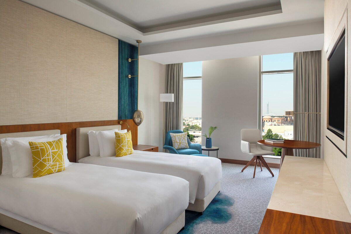 A hotel room with two beds, a desk, a chair, and a view of the city.