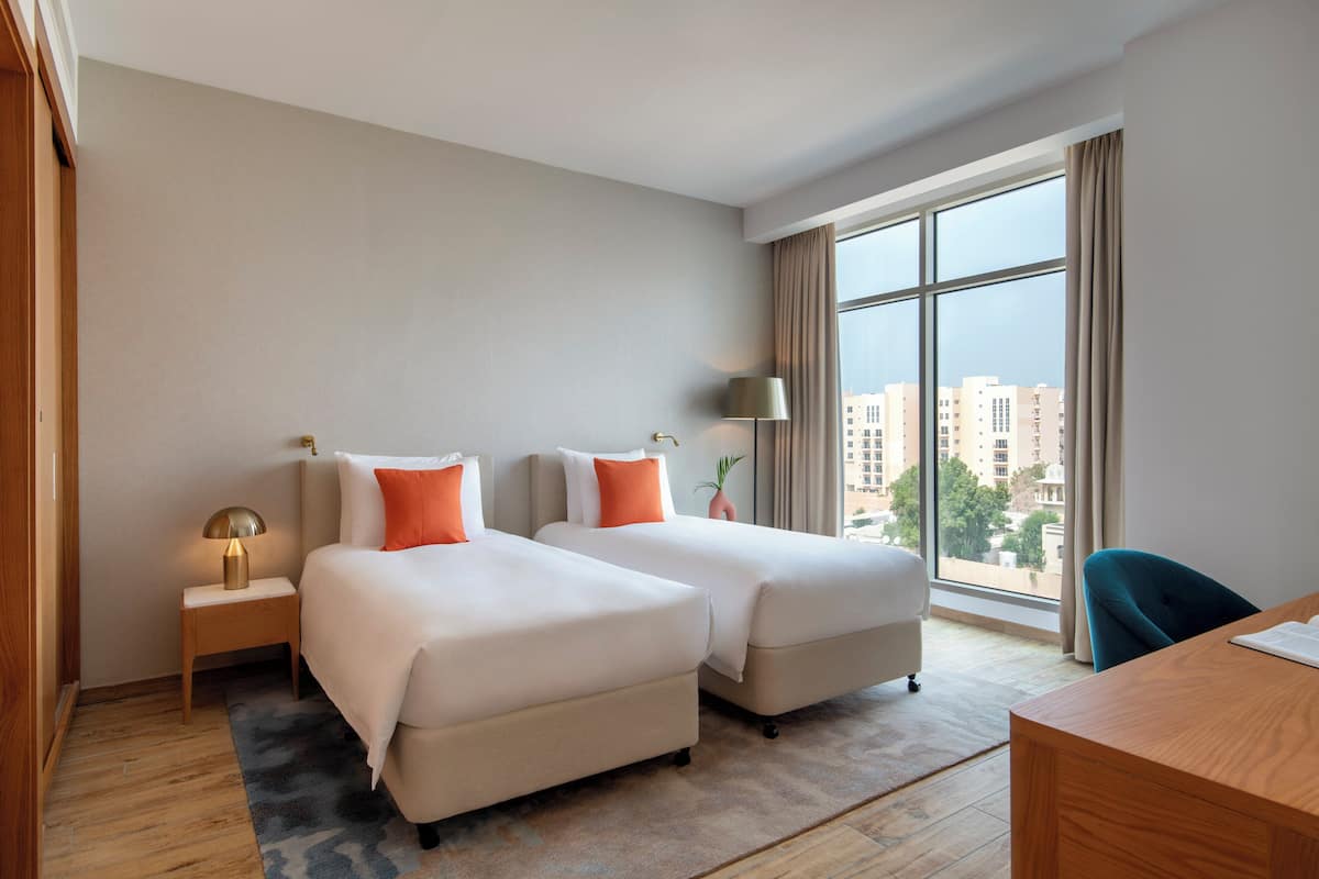 Habitación de hotel con dos camas, un escritorio, una lámpara y vistas a edificios a través de amplias ventanas.