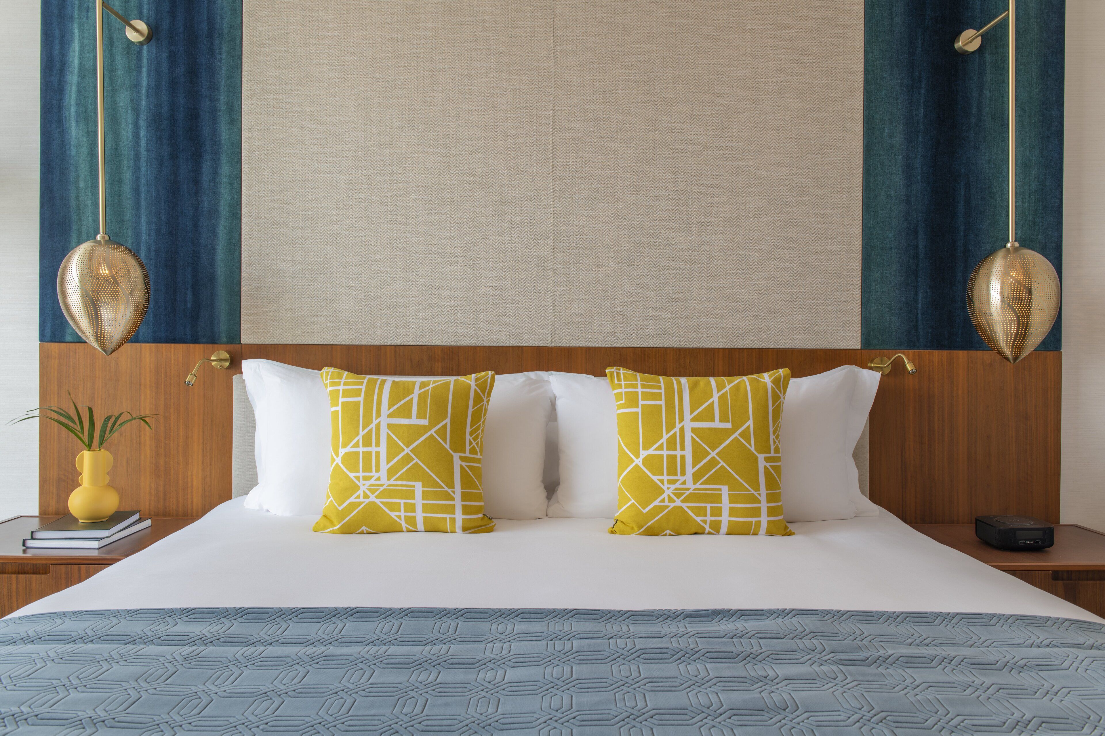 A hotel room with a large bed, two decorative pillows, a bedside table with a book and a vase, and a wall-mounted lamp.
