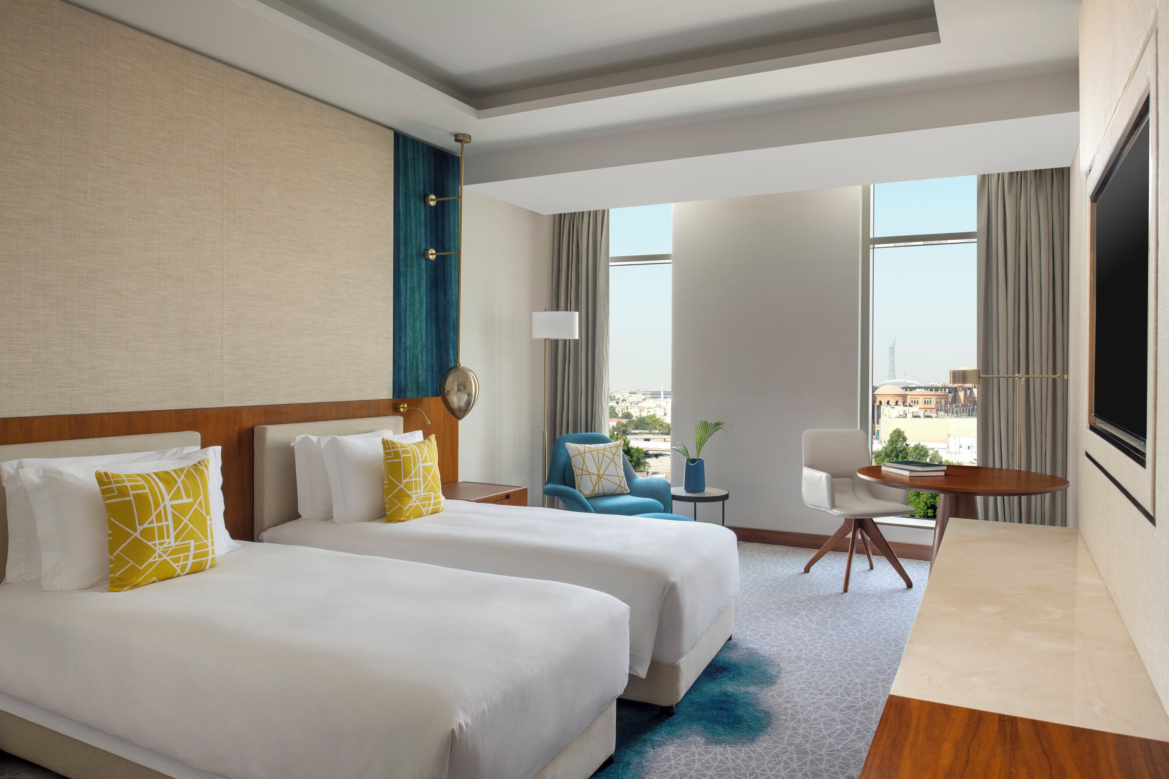 A hotel room with two beds, a desk, a chair, and a view of the city.